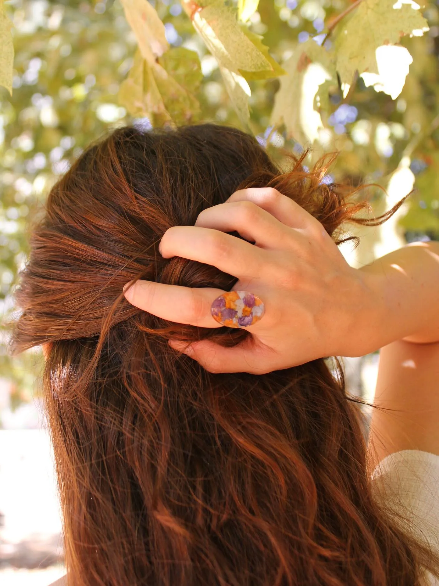Anillo ovalado con pétalos morados y naranjas en una mano recogiéndose el pelo