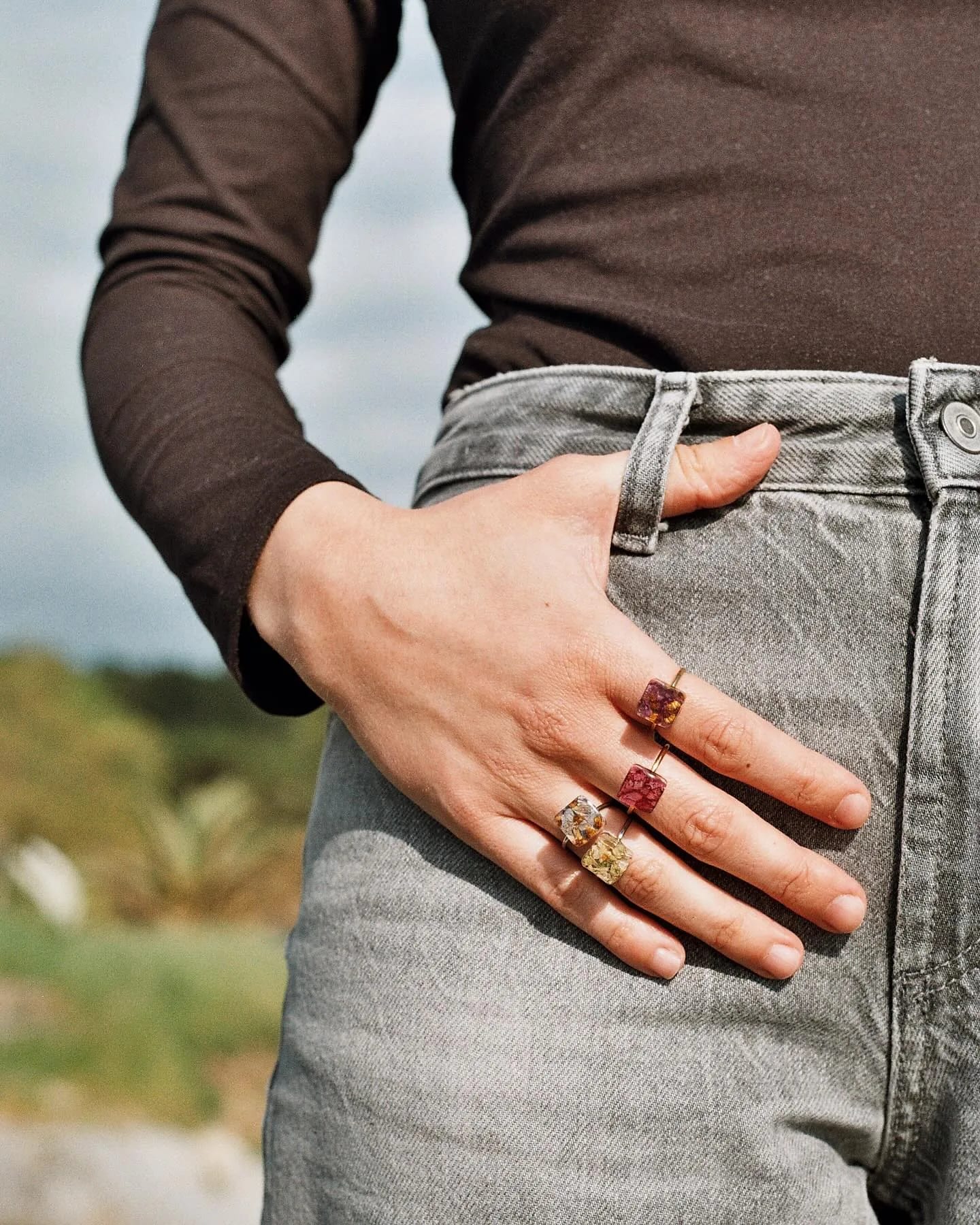 Tres anillos de piedra cuadrada ámbar, rosa y amarilla en una mano apoyada en el bolsillo