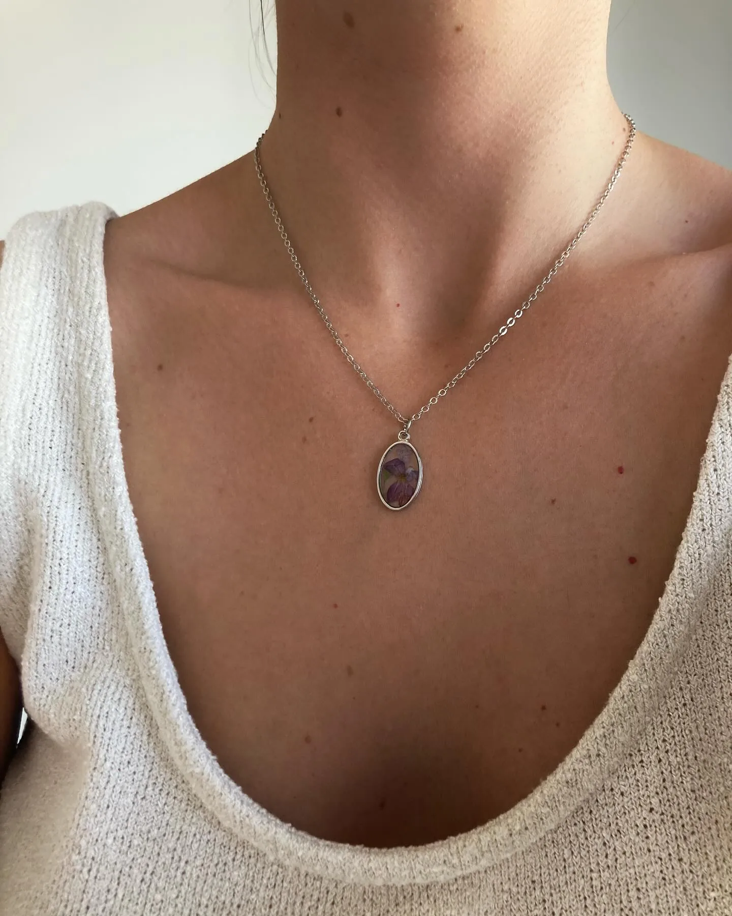 Colgante ovalado plateado con una flor morada puesto al cuello, con jersey claro