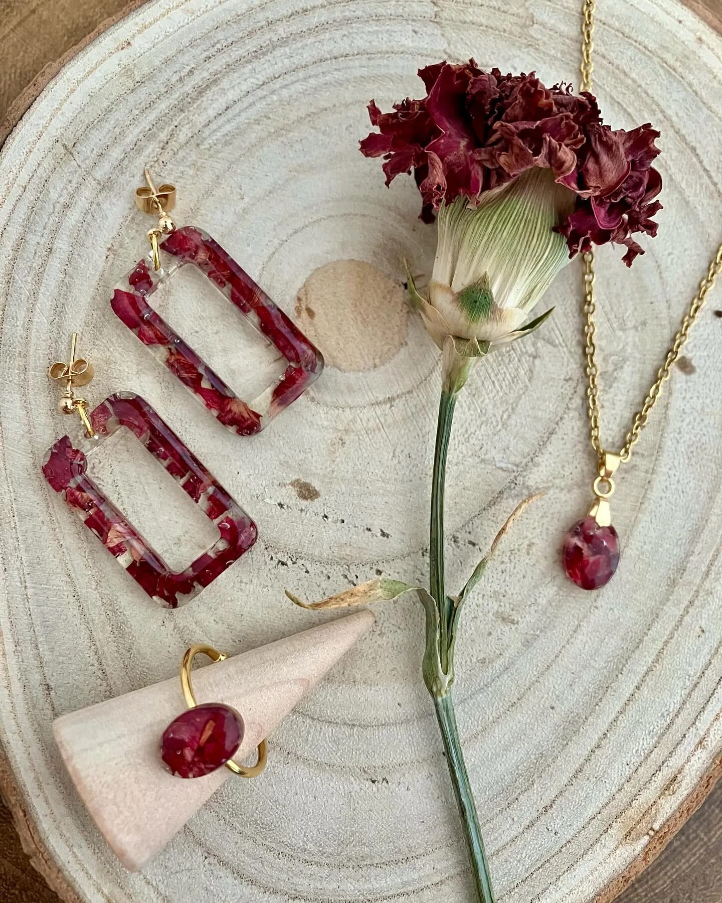 Pendientes, anillo y colgante granates sobre una rodaja de madera, junto a un clavel