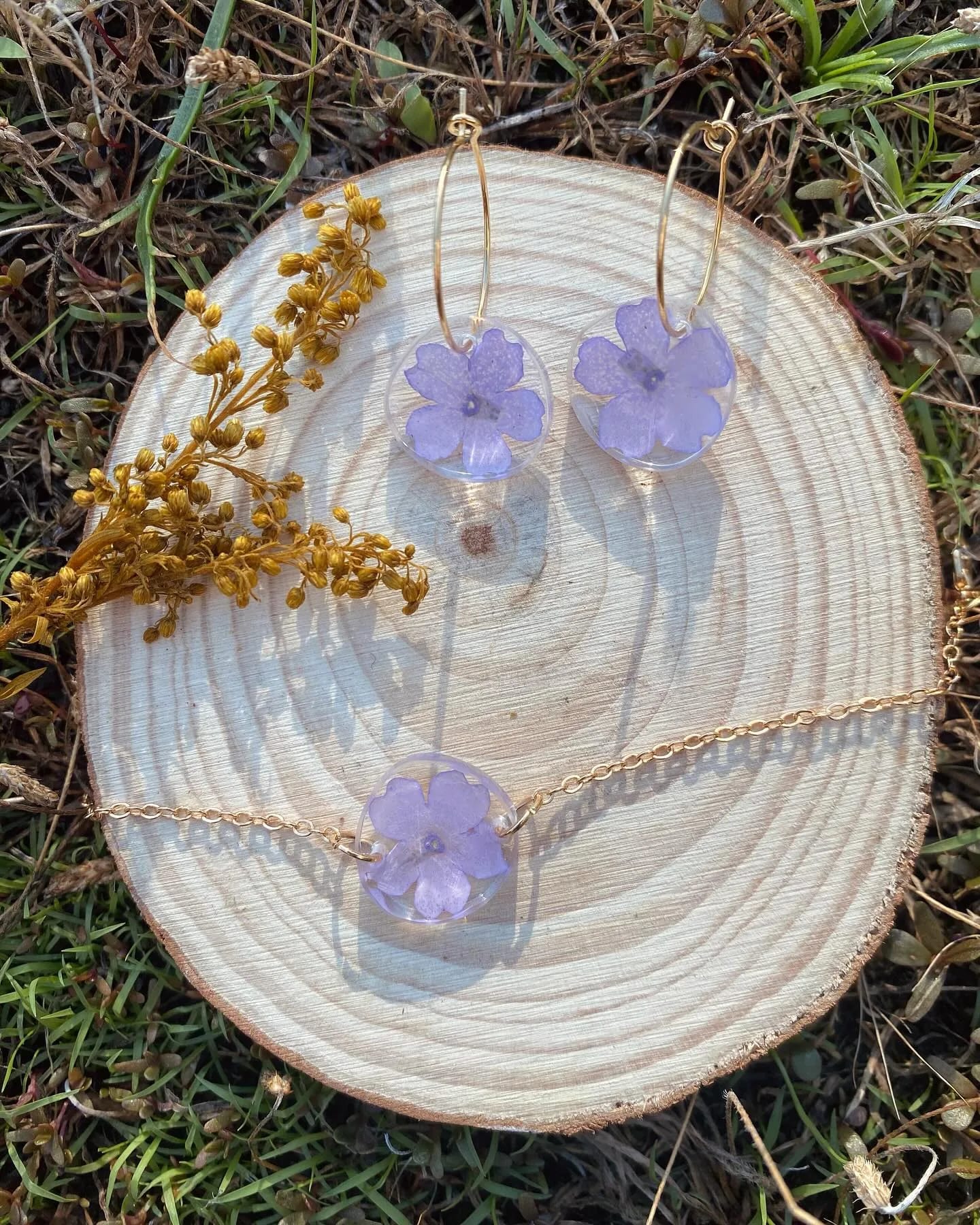 Pendientes de aro y colgante con flores lilas sobre una rodaja de madera, en la hierba