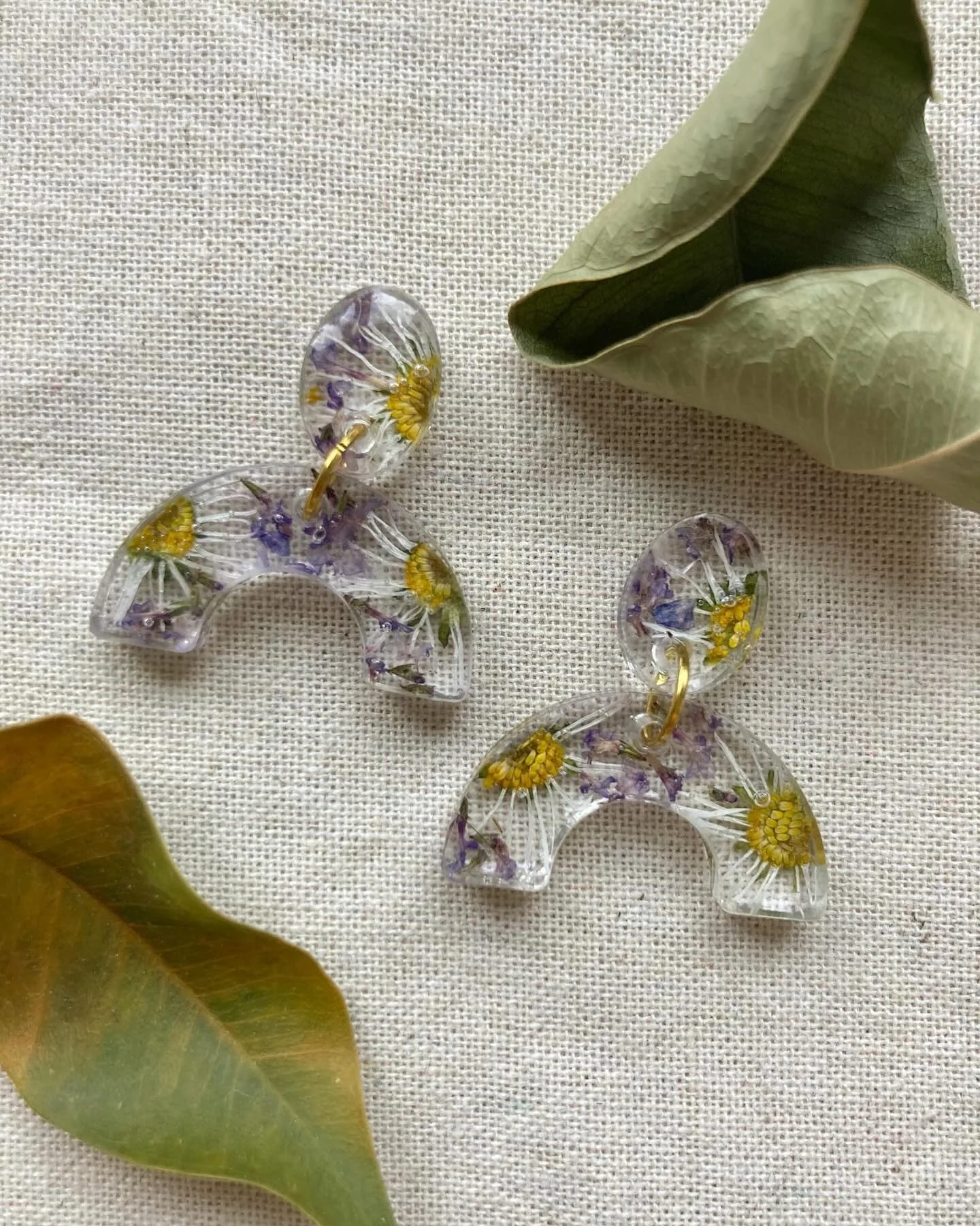 Dos pendientes de arco con margaritas amarillas y flores lilas sobre lino, entre hojas