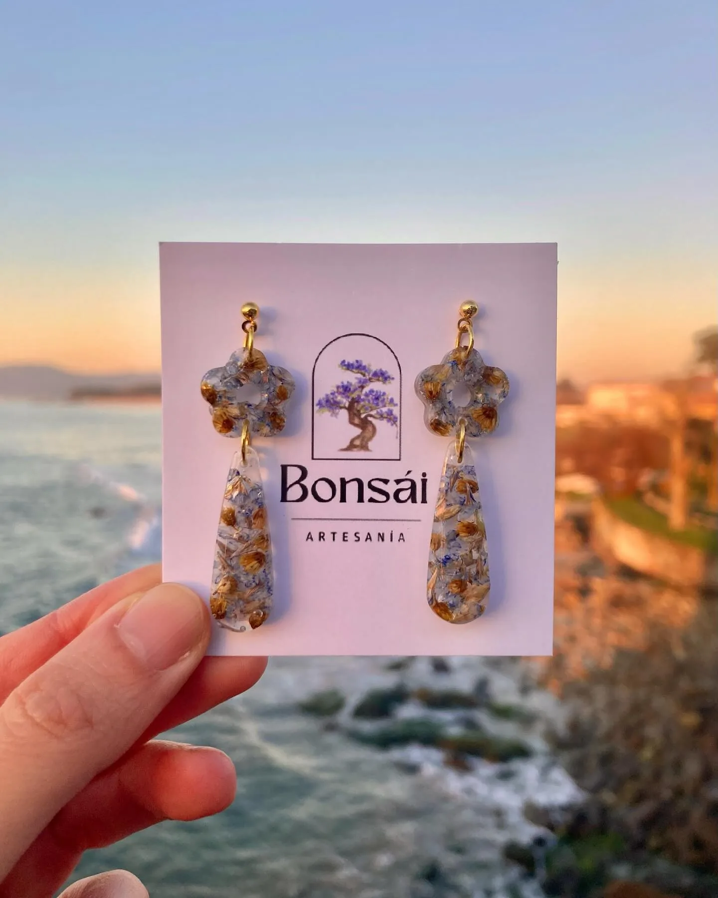 Pendientes de flor y gota en su tarjeta, sostenidos frente al mar al atardecer