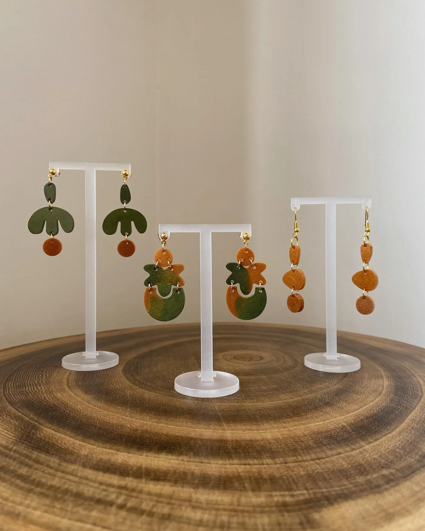 Tres pares de pendientes geométricos verdes y naranjas en expositores sobre madera