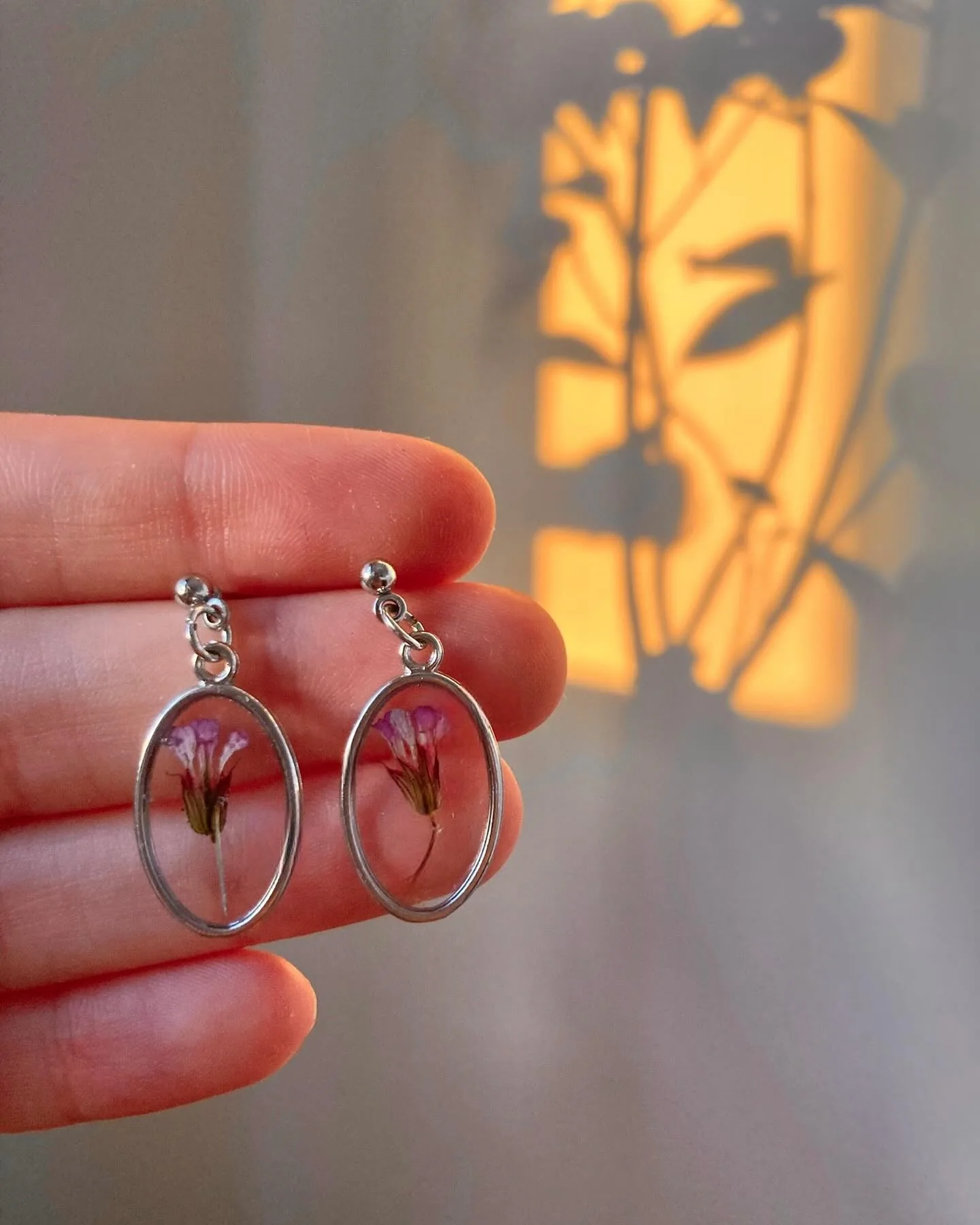 Dos pendientes ovalados plateados con una flor rosa, sostenidos en una mano