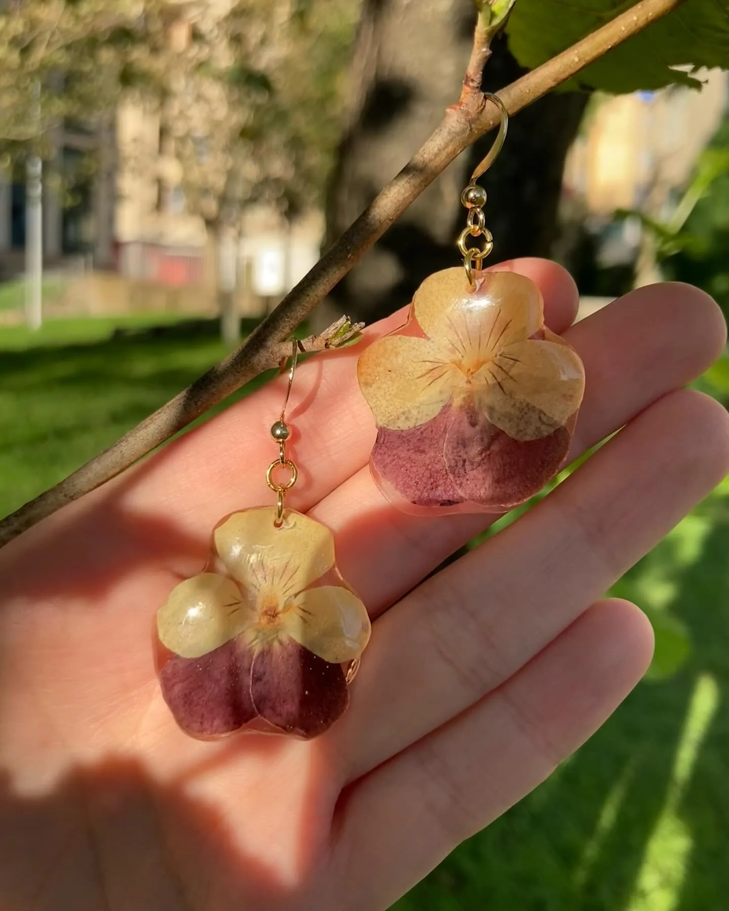 Dos pendientes con flores de pensamiento crema y burdeos sostenidos en una mano al sol