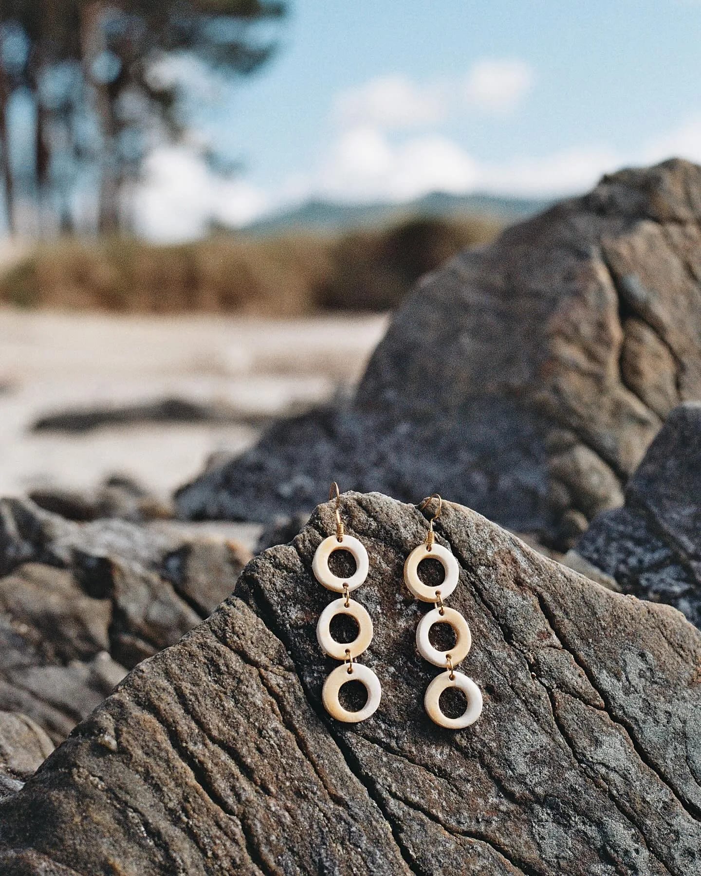 Pendientes de tres aros color crema apoyados en una roca, con la playa al fondo