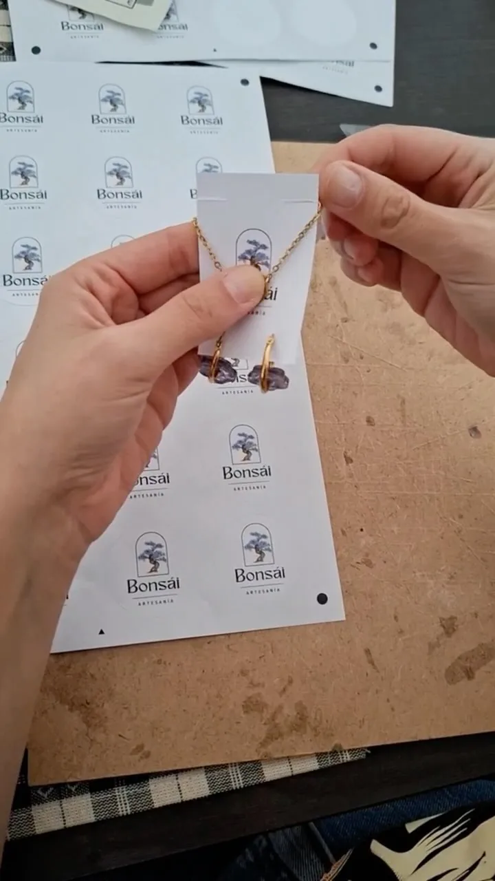 Dos manos montando unos pendientes en una tarjeta con el logo
